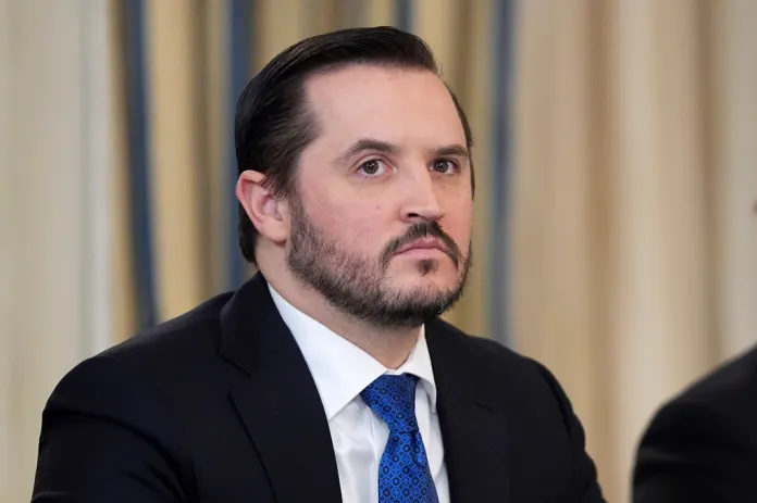 Taylor Budowich, dressed in a suit, looks at something out of frame.