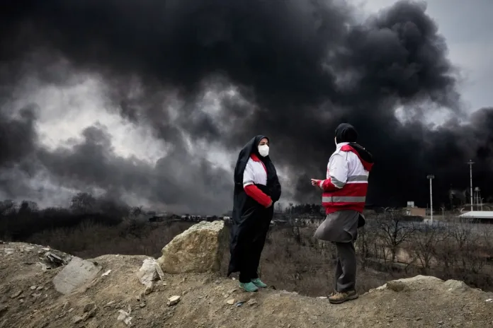 Two women from the Iranian Red Crescent Society stand as a thick plume of smoke from a U.S.-Israeli strike on an oil storage facility late Saturday rises into the sky in Tehran